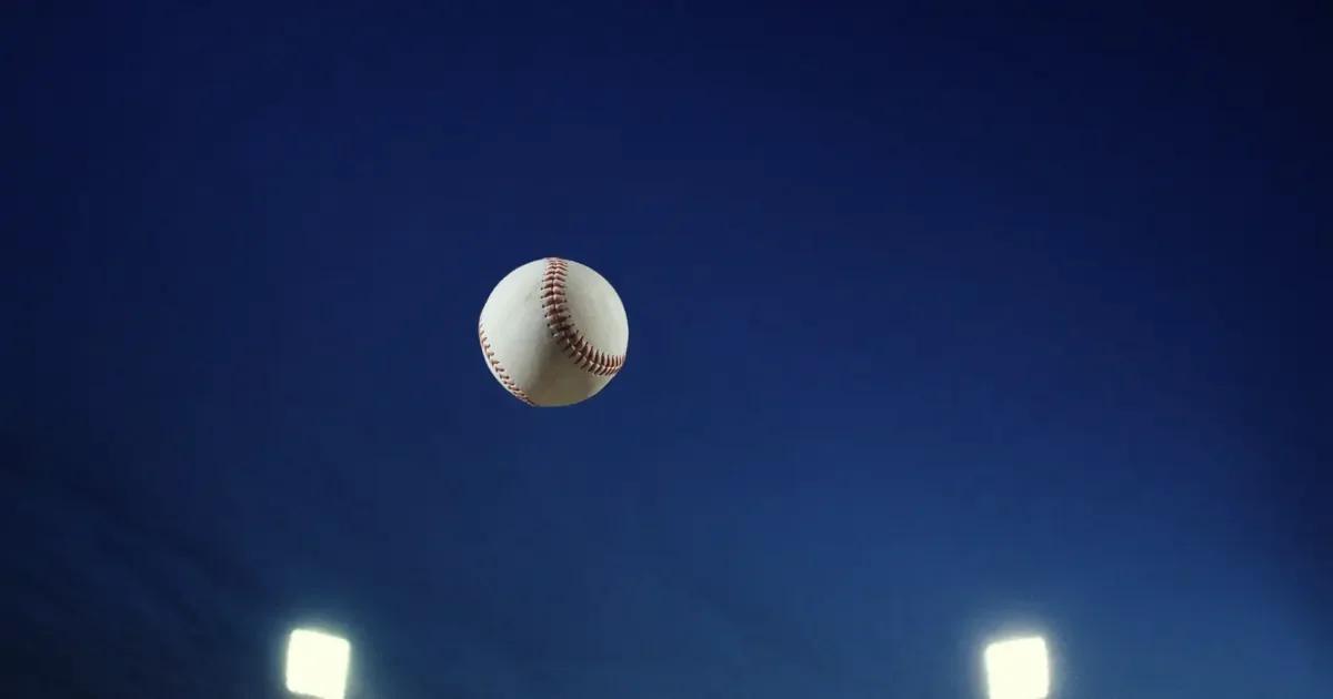 A baseball in mid air against a deep blue evening sky above stadium lights